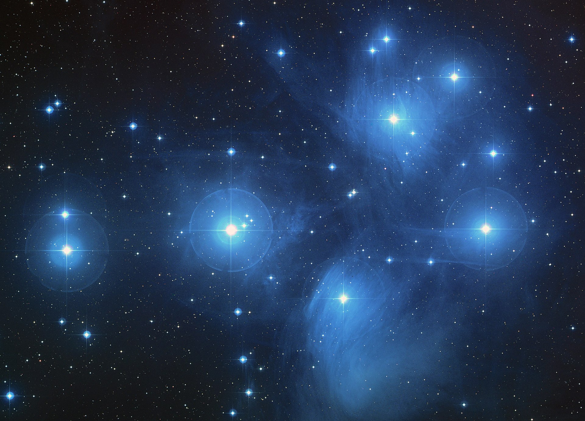 An image of the Pleiades, a loose group of seven bright stars and a smattering of other stars. Each star is a bright blue, and between the stars are wispy tendrils of nebulosity.