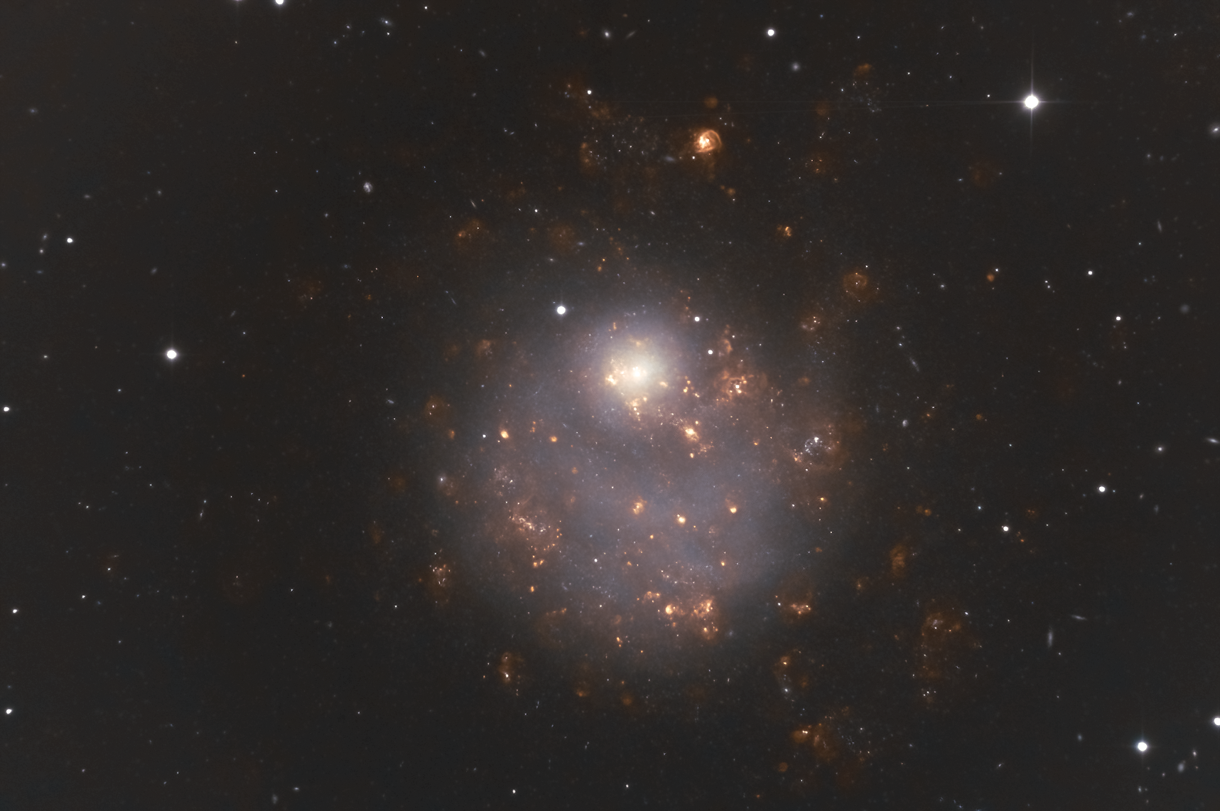A color image of a galaxy. The galaxy shows a faint white featureless disk on top of which are numerous red clumps of light. There is also a featureless white circular bulge towards the top of the faint white disk.